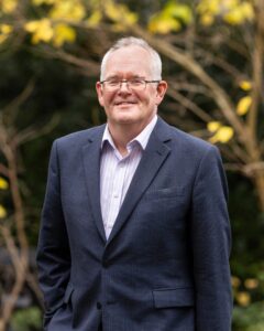 Portrait of Professor Mark Birkin in front of trees - male in blue suit, with glasses, smiles at the camera.
