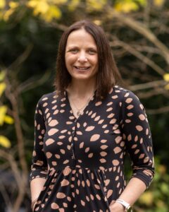 Portrait of Professor Michelle Morris in front of trees - female in black dress with brown leaves, smiles at the camera.