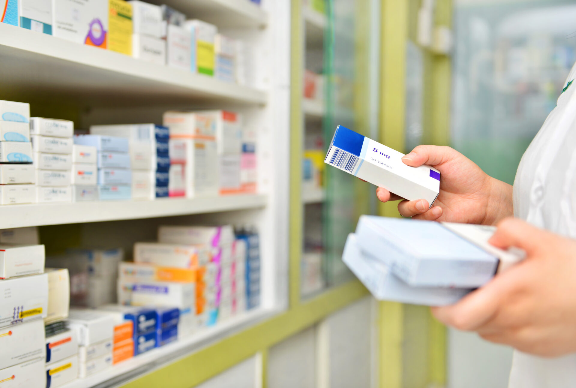 A photograph showing a closeup of a pharmacy shelf stacked with small boxes of medicine; in focus are the hands of a pharmacist picking up a selection of boxes from the shelf.
