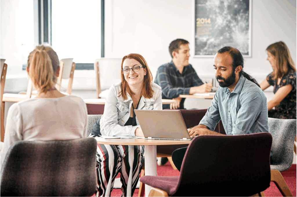 A photograph of a number of people sitting around small tables, talking, smiling, working on laptops, and taking notes.