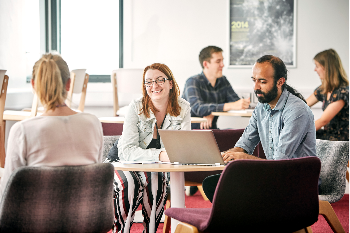 A photograph of a number of people sitting around small tables, talking, smiling, working on laptops, and taking notes.