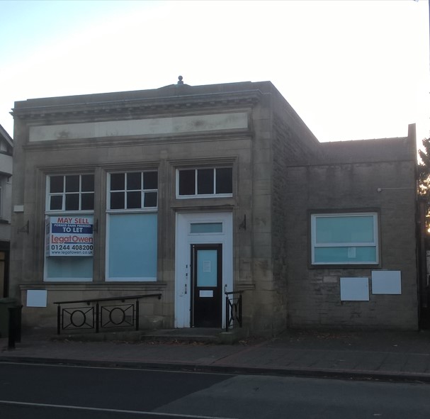 Shows a boarded up bank building, signage on building reads 'To let, may sell'