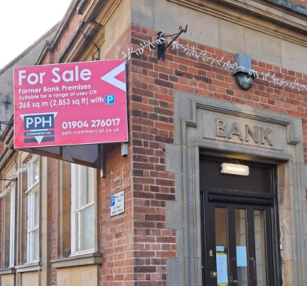 Shows a traditional bank building, wooden door with bank etched in stone above.  Affixed to the side of the building is a vibrant pink 'For Sale' sign 