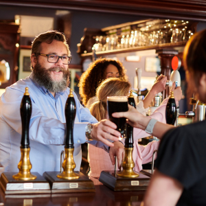 Man with beard and glasses hands over pint of guiness to unidentifiable man, in a traditional British pub