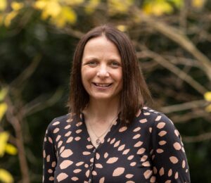 Portrait of Professor Michelle Morris in front of trees - female in black dress with brown leaves, smiles at the camera.