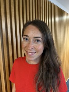 Dr Emma Wilkins, female academic, wearing a red top, looks towards camera and smiles.