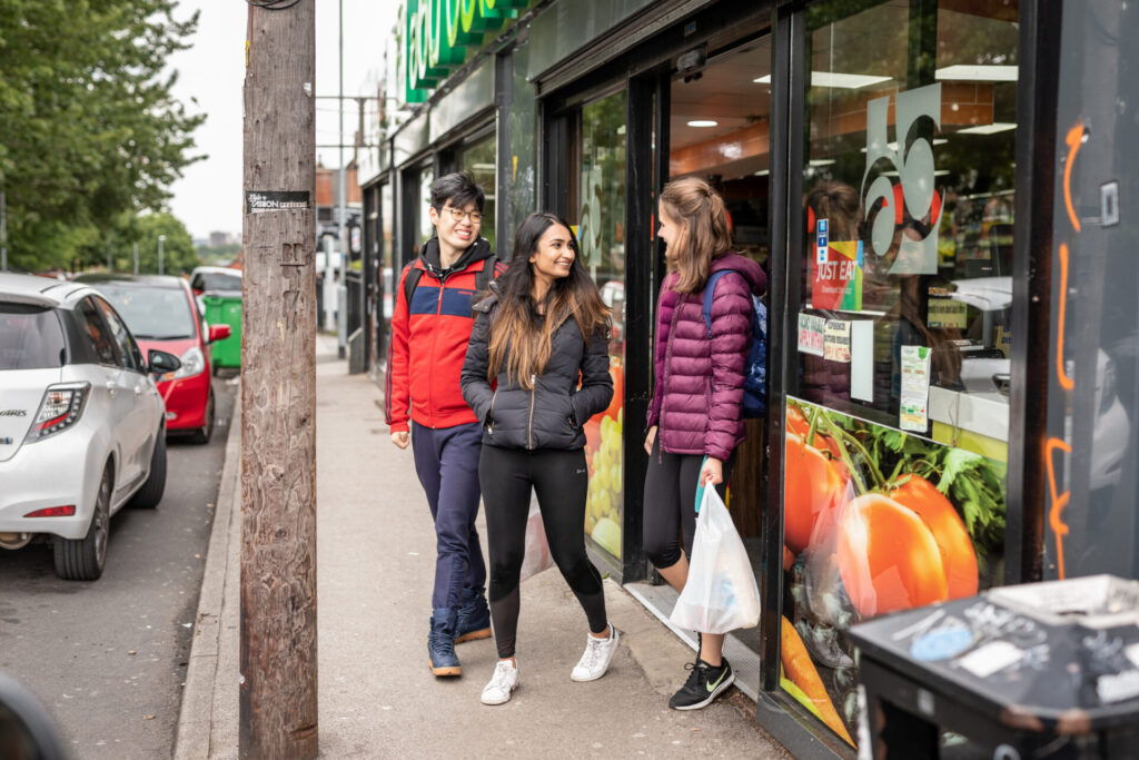 Two females and a male exiting a convenience style store on to the pavement. All three wear winter jackets and one carries a plastic bag.