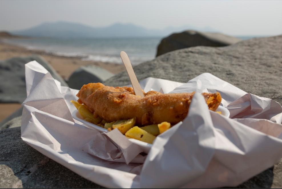 Fish and chips wrapped in paper in the foreground, with a beach scene in the background